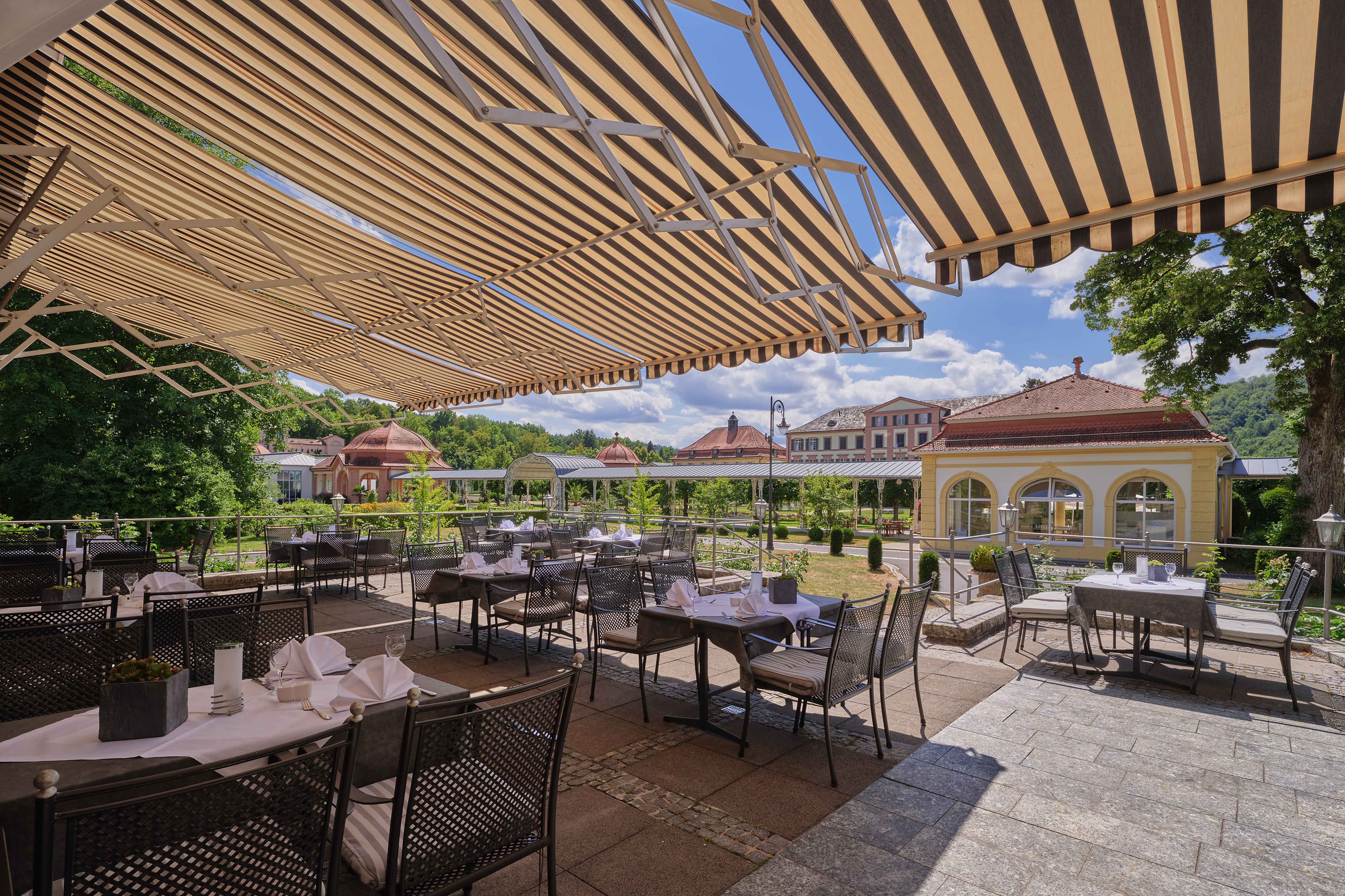 Das Bild zeigt eine einladende Außenterrasse eines Restaurants oder Cafés mit Blick auf einen gepflegten Park und historische Gebäude.